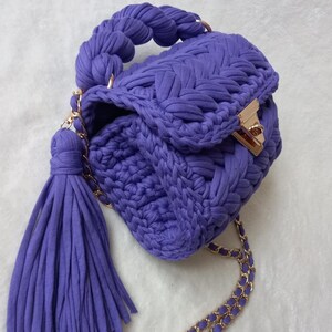 May include: A small, purple crocheted handbag with a braided handle and a gold chain strap. The bag has a tassel and a gold clasp.