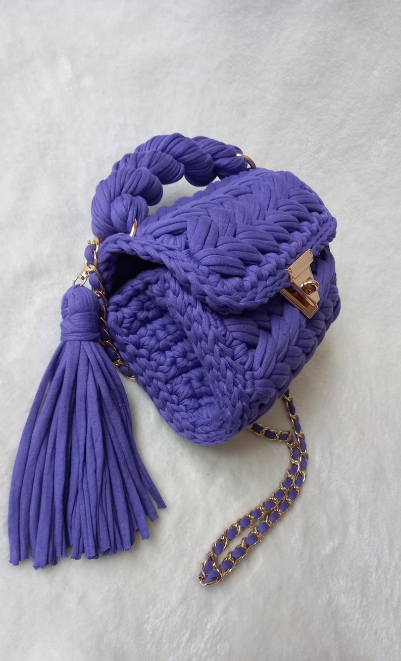 May include: A small, purple crocheted handbag with a braided handle and a gold chain strap. The bag has a tassel and a gold clasp.