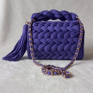 May include: A small purple knitted handbag with a gold chain strap and a tassel. The bag has a braided handle and a woven design.
