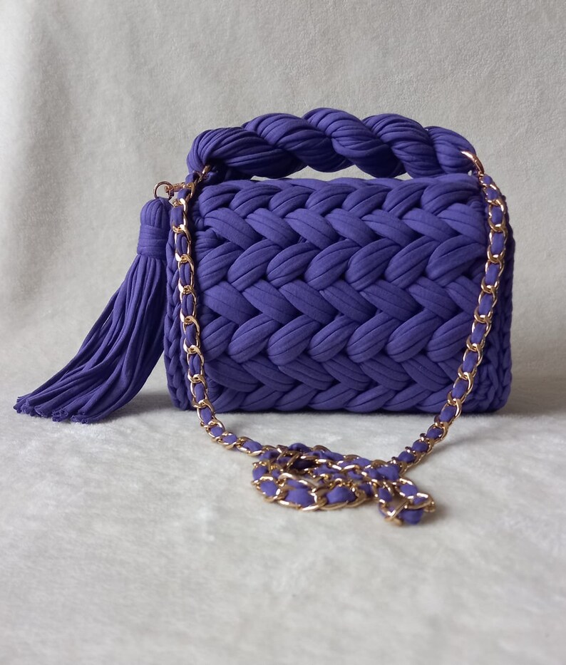 May include: A small purple knitted handbag with a gold chain strap and a tassel. The bag has a braided handle and a woven design.
