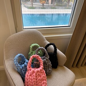 Puede incluir: Cinco bolsas de mano de punto en diferentes colores, incluyendo rosa, azul, verde, gris y negro, est&aacute;n dispuestas en un sill&oacute;n gris claro. Las bolsas tienen un dise&ntilde;o trenzado y est&aacute;n hechas de hilo suave.