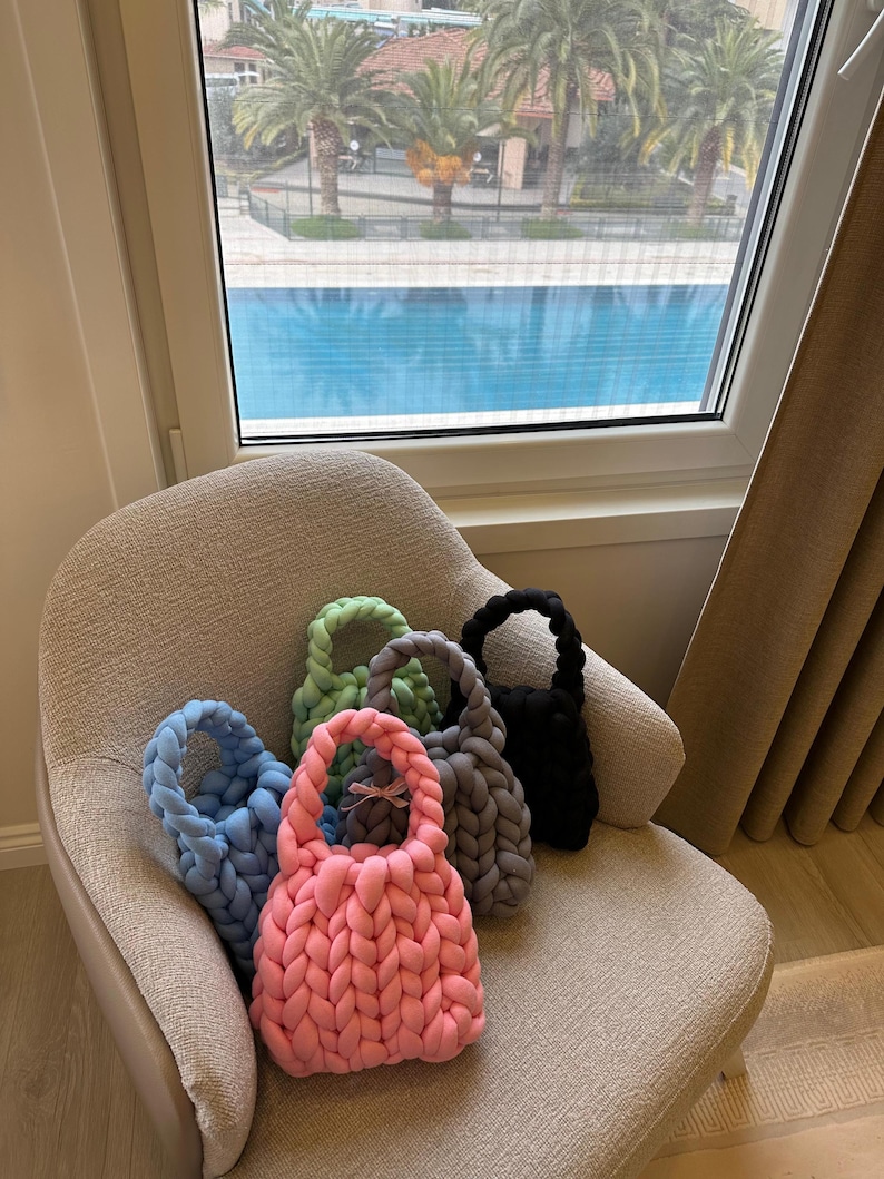 Puede incluir: Cinco bolsas de mano de punto en diferentes colores, incluyendo rosa, azul, verde, gris y negro, est&aacute;n dispuestas en un sill&oacute;n gris claro. Las bolsas tienen un dise&ntilde;o trenzado y est&aacute;n hechas de hilo suave.
