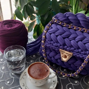 May include: A purple crocheted bag with a gold chain strap, a cup of coffee, and a glass of water on a table with a black and white patterned tablecloth.