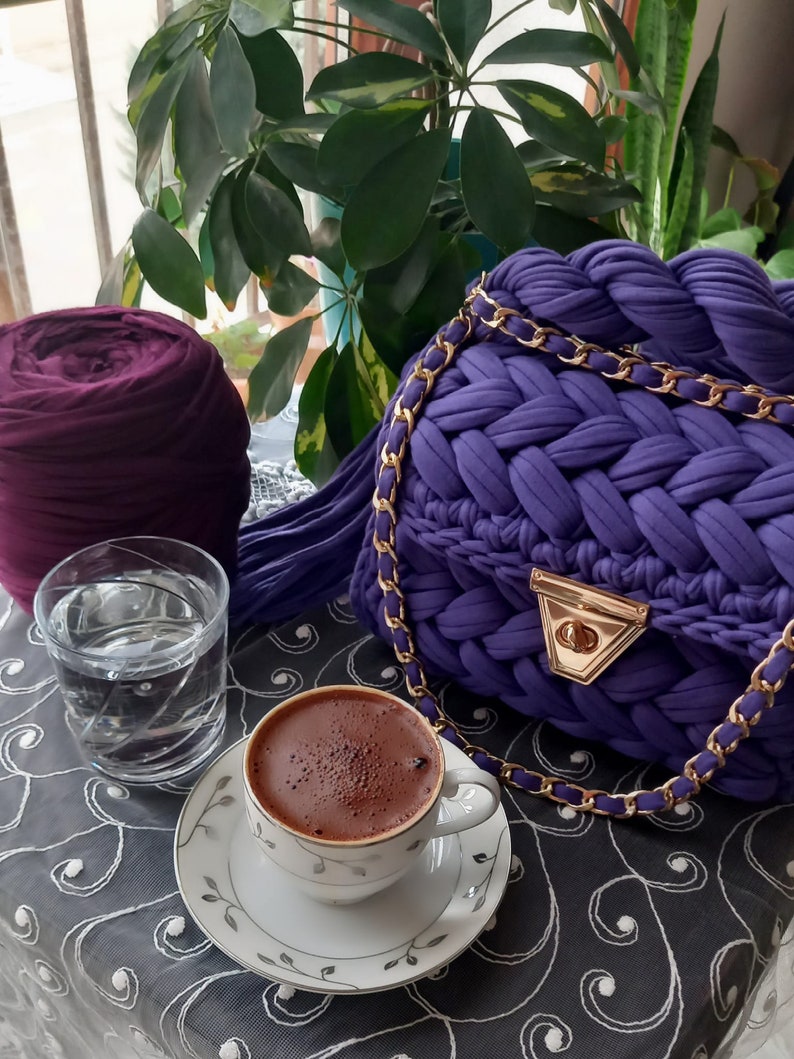 May include: A purple crocheted bag with a gold chain strap, a cup of coffee, and a glass of water on a table with a black and white patterned tablecloth.