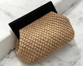 Könnte beinhalten: Eine geflochtene Strohtasche mit schwarzem Klappenverschluss. Die Tasche liegt auf einer weißen Marmorfläche.