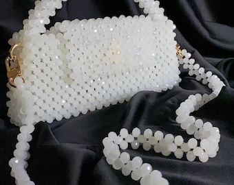 May include: A white beaded handbag with a matching beaded shoulder strap and handle. The rectangular bag has a gold-colored clasp and is set against a black background. The beads are small and faceted, giving the bag a sparkling appearance.