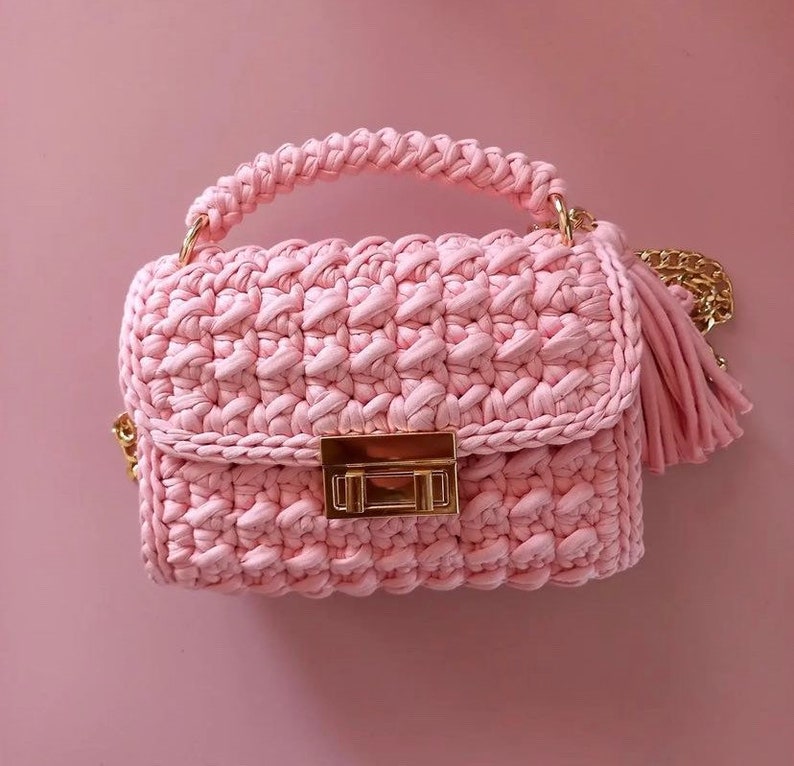 May include: A pink crocheted handbag with a gold chain strap and a gold clasp. The bag has a tassel on the strap.