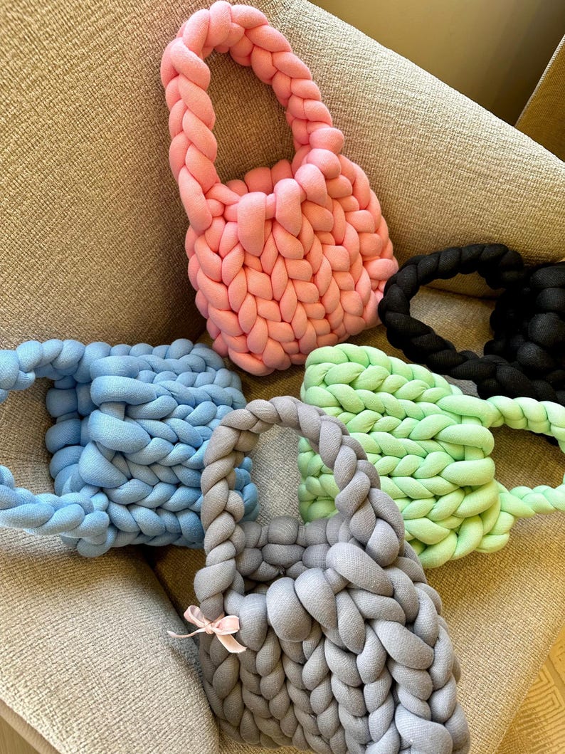 Puede incluir: Cinco bolsas de mano de punto en diferentes colores: rosa, azul claro, verde claro, gris y negro. Las bolsas est&aacute;n hechas de hilo grueso y tienen un asa trenzada.