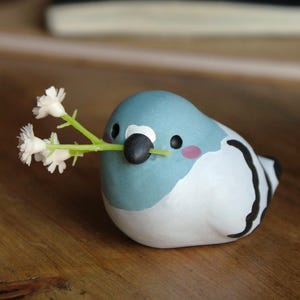 Può includere: Figurina di uccello in ceramica con testa blu, corpo bianco e dettagli neri. L'uccello tiene un rametto di fiori bianchi nel becco. La figurina ha guance rosa ed è appoggiata su una superficie di legno.