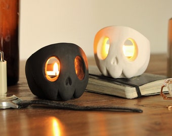 May include: Two ceramic skull candle holders, one black and one white, with lit candles inside. The holders are on a wooden surface with a book and glasses.