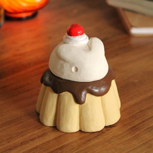 Puede incluir: Una figurita de cerámica de un postre de pudín con una cobertura de crema batida blanca y una cereza roja en la parte superior. El pudín está decorado con salsa de chocolate marrón.