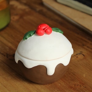 Op de afbeelding: Een keramische sieradendoos in de vorm van een kerstpudding. De basis is bruin, met een witte bovenkant en een rode kers en groene bladdecoratie. De doos staat op een houten oppervlak.