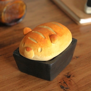 Könnte beinhalten: Ein Keramikbehälter in Brotform mit einem Tigergesichtsdesign. Der Behälter befindet sich in einer schwarzen quadratischen Schale.