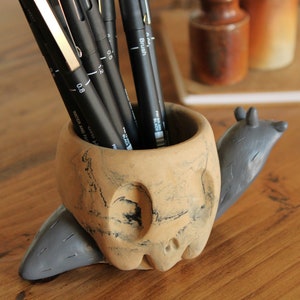 May include: A ceramic snail-shaped pencil holder with a textured, brown and gray finish. The holder is filled with black pens.
