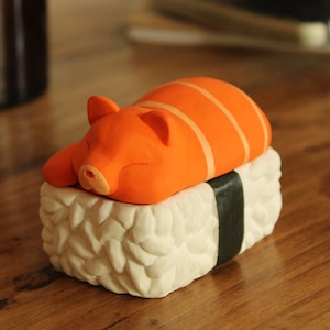 May include: A small orange pig figurine shaped like a piece of sushi, with white rice and a black seaweed wrap.