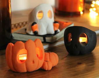 May include: Three Halloween themed candle holders: a white ghost, a black skull, and an orange pumpkin. Each holder has a small opening for a tea light candle.