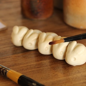May include: A set of five white ceramic hand-shaped figurines being painted with a brown paintbrush.