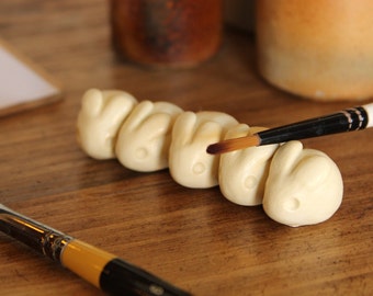 May include: A set of five white ceramic hand-shaped figurines being painted with a brown paintbrush.