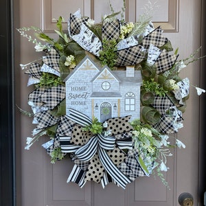 May include: A black and white checkered and polka dot ribbon bow with a house-shaped sign that reads "Home Sweet Home" on a green and brown wreath with white flowers.