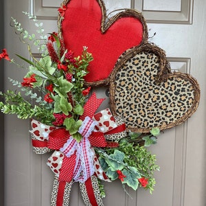 May include: A Valentine's Day wreath with two hearts, one red and one with a leopard print. The wreath is decorated with a red and white bow with a white lace accent and greenery.