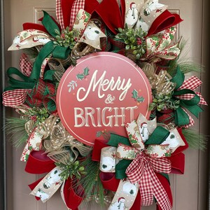 May include: A Christmas wreath with red, green, and white ribbon, burlap, and greenery. The wreath has a red sign that says "Merry & Bright".