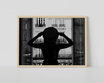 Op de afbeelding: Zwart-wit ingelijste print van een silhouet van een persoon met een hoed, die uit een raam kijkt. De armen van de persoon zijn omhoog, de handen raken de hoed aan. De achtergrond toont een stadsgezicht met gebouwen.