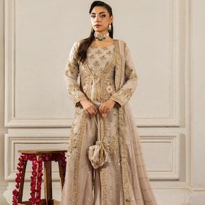 Puede incluir: Un vestido formal beige con bordados y adornos dorados. El vestido presenta una chaqueta larga, una bufanda a juego y un pequeño bolso. El atuendo se usa con un collarín y aretes verdes. El fondo incluye una alfombra roja y un pequeño taburete de madera.