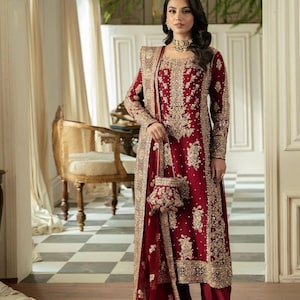 May include: A burgundy formal ensemble featuring intricate gold embroidery. The outfit includes a long-sleeved tunic, matching palazzo pants, a long jacket, and a small purse. The ensemble is worn with a gold choker necklace.