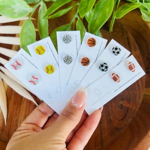 May include: A collection of sports-themed stud earrings, including baseball, softball, volleyball, basketball, soccer, and football designs. Each earring is displayed on a white card with the website address www.mandihomemadecreations.com.