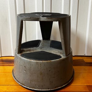 May include: A gray metal step stool with a round top and a lower, wider base. The top and the three step pads have black, non-slip surfaces. The stool has a sturdy, industrial design, suitable for reaching higher shelves or tasks.