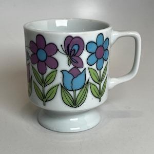 May include: White ceramic mug with a pedestal base, adorned with a floral pattern. The design features purple, blue, and green flowers and a butterfly. The mug has a handle and is suitable for hot beverages.