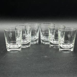 May include: A set of six clear glass shot glasses, each featuring a delicate etched starburst pattern on the exterior. The glasses have a cylindrical shape with a thick, solid base and smooth, transparent walls, arranged in a cluster.