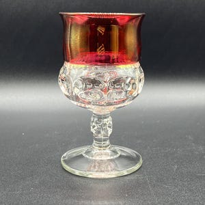 May include: A vintage clear glass goblet with a red rim and a decorative, patterned base. The goblet has a round foot and a detailed stem. The red rim has a gold-colored design. The glass is transparent, allowing light to pass through.