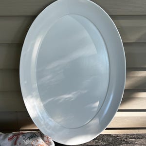 May include: A large, plain white oval serving platter with a smooth, glossy finish. The platter features a wide, flat rim and a deep, recessed center area. The surface reflects light, creating subtle highlights across the white ceramic material.