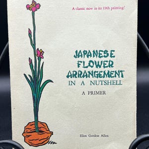 May include: A book titled "Japanese Flower Arrangement in a Nutshell, A Primer" is displayed. The cover features a botanical illustration of a flower with pink blossoms and green leaves. The text "A classic now in its 19th printing!" is also visible.