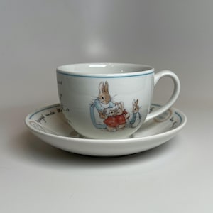 May include: A white ceramic teacup and saucer set featuring a blue rim and illustrations of Peter Rabbit characters. The cup displays a group of rabbits in blue and red clothing, while the saucer includes cursive black text along the edge.