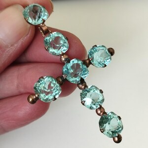 May include: A vintage cross pendant featuring seven light blue, faceted gemstones. The gemstones are set in a copper-toned metal setting. The cross is held by a hand against a neutral background. The gemstones are round and have a slight sparkle.