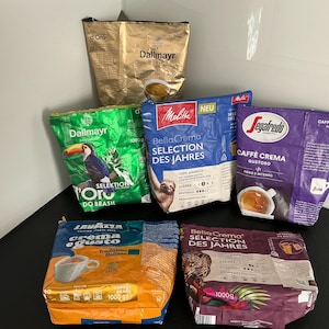 Könnte beinhalten: Verschiedene Kaffeebohnenpackungen in verschiedenen Farben, darunter Gold, Grün, Blau, Lila und Orange. Die Packungen tragen Markennamen wie Dallmayr, Melitta, Lavazza und Segafredo, mit Text und Bildern von Kaffeetassen und Bohnen.
