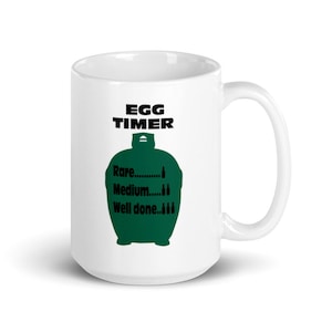 Puede incluir: Taza de cerámica blanca con asa negra. La taza tiene el texto "EGG TIMER" sobre un gráfico verde. El gráfico incluye las palabras "Rare", "Medium" y "Well done", con ilustraciones de huevos.
