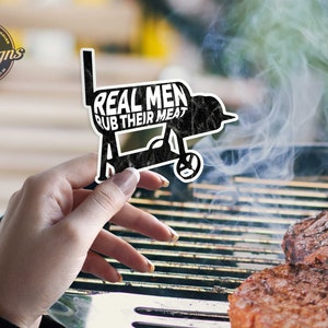 Puede incluir: Una pegatina en blanco y negro de una parrilla con el texto "Real Men Rub Their Meat". La pegatina se sostiene sobre una parrilla con dos hamburguesas cocinándose.