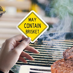 Puede incluir: Un cartel amarillo en forma de diamante con texto negro que dice "Puede contener brisket". El cartel se sostiene frente a una parrilla con humo saliendo de ella.