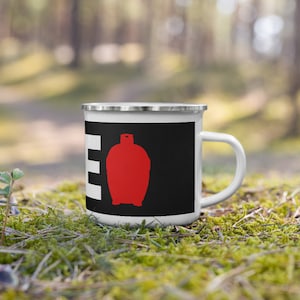 Puede incluir: Taza de esmalte blanco con borde y asa plateados. La taza presenta un panel negro con un gráfico rojo y la letra blanca "E". La taza está sobre musgo verde.