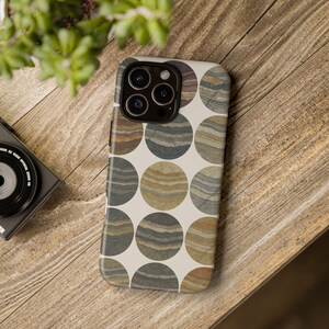 May include: A smartphone case featuring a repeating pattern of layered, circular designs in shades of brown, tan, and gray. The case is set against a light background and has a camera cutout. The case is on a wooden surface.