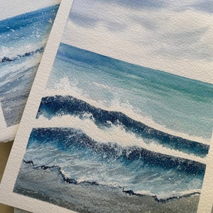 May include: A watercolour painting of a seascape with blue and white waves crashing on a sandy beach. The sky is a light blue with white clouds.