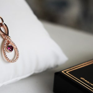 May include: A rose gold infinity necklace with a small red heart pendant and clear cubic zirconia stones.