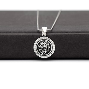 May include: A silver necklace with a round pendant featuring Arabic calligraphy in black on a white background. The pendant has a decorative border.