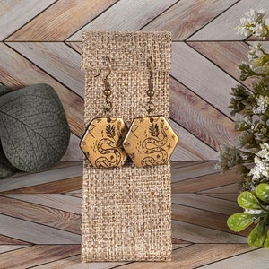 May include: A pair of hexagon-shaped earrings with a gold finish. Each earring features a black snake and botanical design. The earrings hang from bronze-colored ear wires and are displayed on a tan fabric stand.