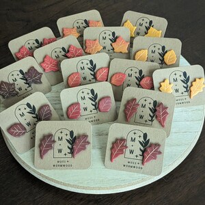 Puede incluir: Pendientes con forma de hoja en colores otoñales, rojo, naranja y amarillo. Cada par se presenta en una pequeña tarjeta cuadrada con el texto "MOSS + WORMWOOD" y un logotipo. Los pendientes están dispuestos sobre una bandeja redonda de madera clara.