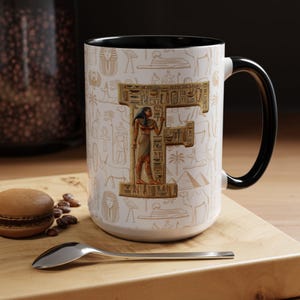 May include: White ceramic mug with a black handle and rim, featuring an Egyptian-themed design. The design includes hieroglyphs and an image of an Egyptian figure. The mug is on a wooden surface.