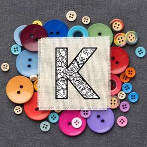 May include: A white fabric with a black and white patterned letter K surrounded by colourful buttons. The buttons are in various shapes and sizes.